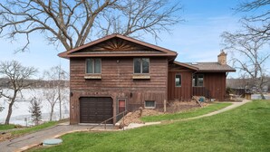 A two-story wooden house with a garage, a covered porch, and a view of a lake. - Cedar Lake getaway - sauna, paddle boards & raft! (Annandale)