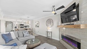 En moderne stue med et fladskærms-tv, en loftsvifte, en pejs og et spiseområde i baggrunden.