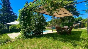 Een overdekt terras met een tafel en stoelen, omringd door weelderige begroeiing en een zwembad op de achtergrond.