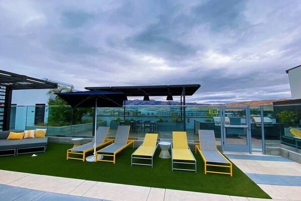A rooftop lounge area with multiple sun loungers, a glass enclosure, and a view of distant mountains. - Modern Luxe Apartment in Heart of Silicon Valley (Milpitas)