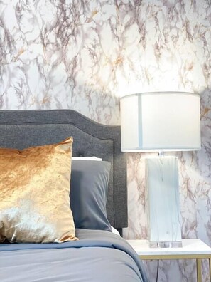 A bed with a dark headboard, a gold and gray pillow, and a white lamp on a bedside table against a marble-patterned wall.