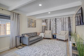 Ein modernes Wohnzimmer mit einem grauen Sofa, zwei Sesseln, einem gemusterten Teppich und einem großen Fenster mit Vorhängen.