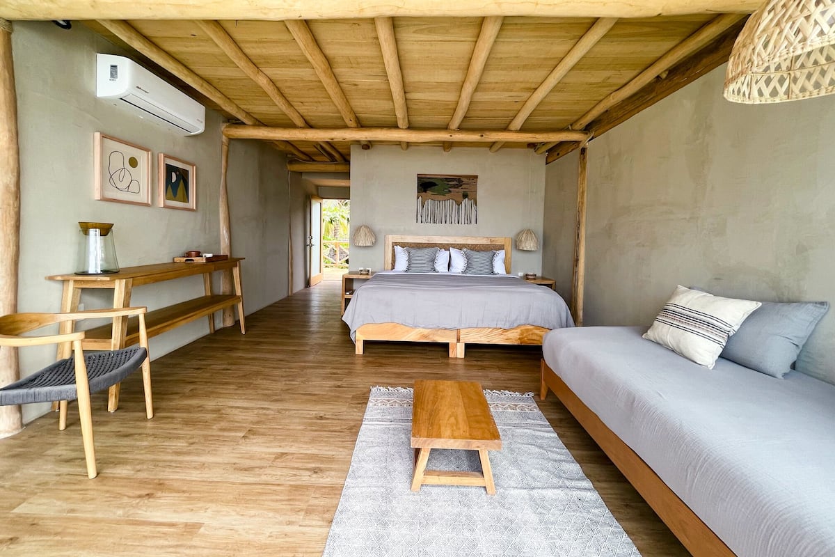 A cozy room with a wooden ceiling, a bed with a grey bedspread, a wooden bench, a small table, and a hanging light fixture.