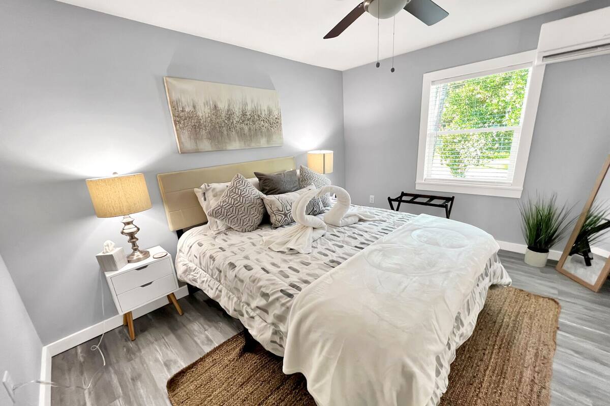 A modern bedroom with a bed, bedside lamps, a ceiling fan, a window with a view of greenery, and a painting on the wall.