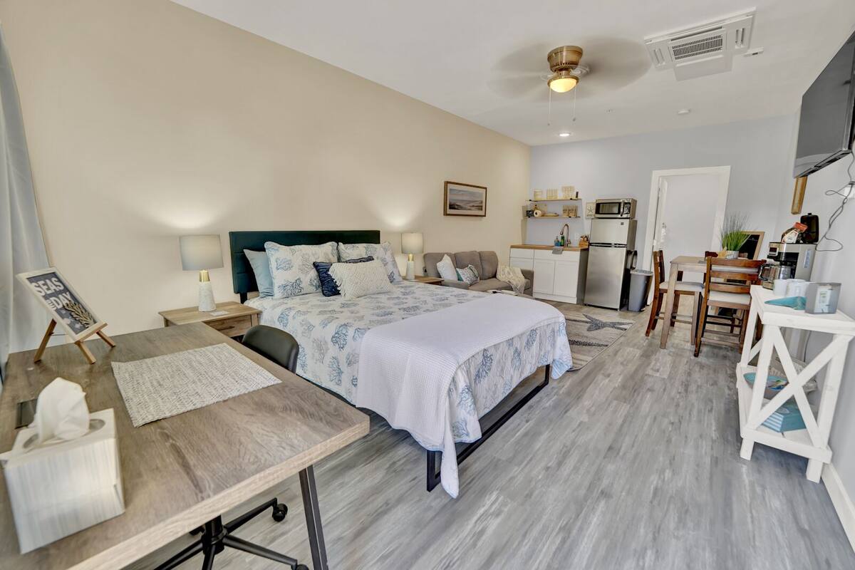 A modern bedroom with a large bed, bedside tables, a desk, and a kitchenette.