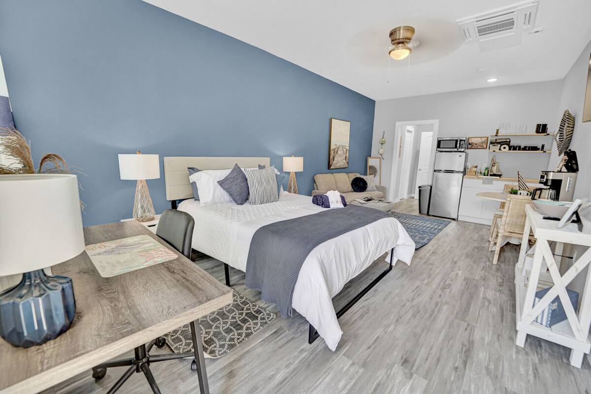 A modern bedroom with a large bed, bedside tables, a desk, and a kitchenette.