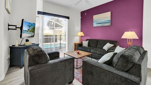 A living room with a purple accent wall, a ceiling fan, a TV mounted on the wall, and a view of the outdoors through a sliding glass door.