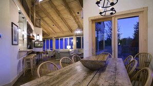 Un comedor amplio con una larga mesa de madera y sillas, un centro de mesa con un tazón grande, y vista al exterior a través de un vidrio.
