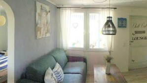 A cozy living room with a sofa, a coffee table, and a TV. There is a door labeled "Lounge" and a window with curtains. - Small oasis of peace surrounded by greenery - 3 minutes from Kromlau Rhododendron Park (Gablenz)