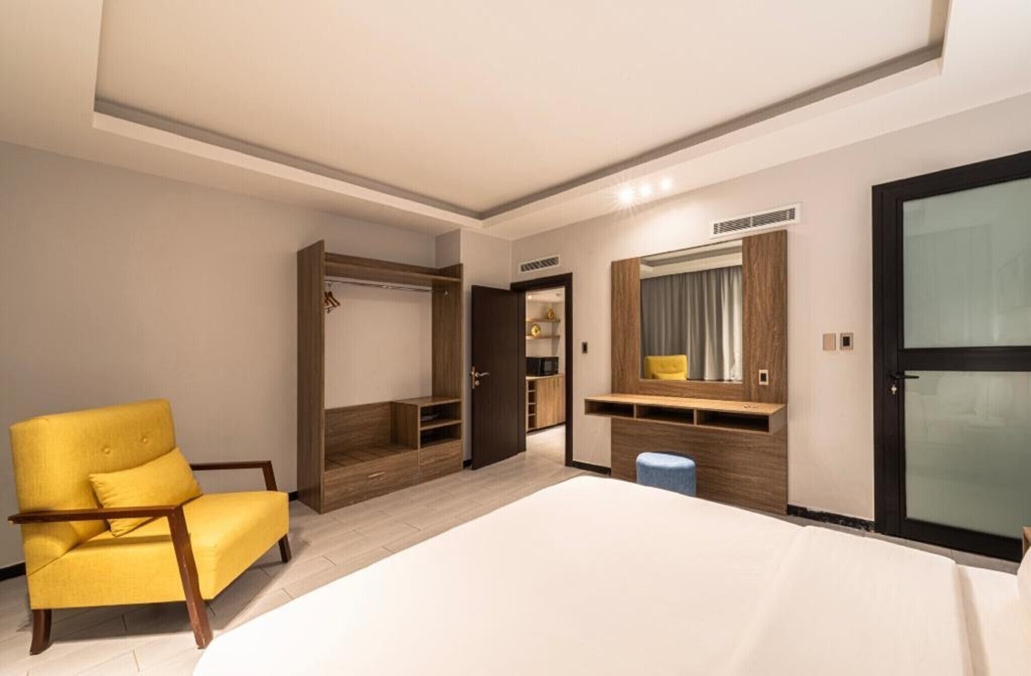Una camera d'albergo con un letto, una poltrona gialla, un armadio, una toeletta con uno specchio e uno sgabello.