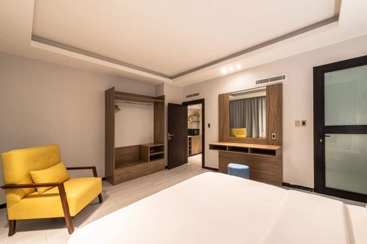 Una habitación de hotel con una cama, un sillón amarillo, un armario, una tocador con espejo y un taburete pequeño.