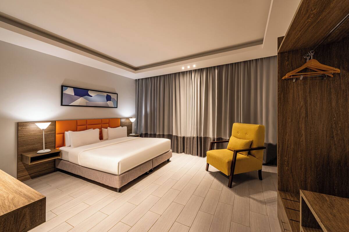 Una habitación de hotel moderna con una cama grande, una mesita de noche con lámpara, una silla y un panel de pared de madera.