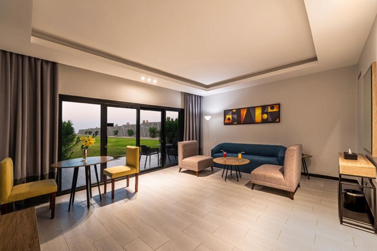 A modern hotel room with a large window, a sofa, a coffee table, and a dining area.