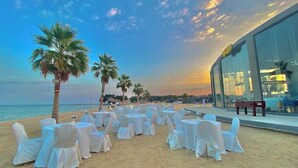 Montaje para un evento en la playa con mesas y sillas redondas, palmeras y un edificio moderno al fondo.