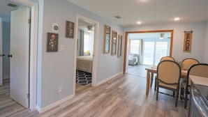 Um quarto espaçoso com soalho em madeira, uma área de jantar com mesa e cadeiras, e uma porta que dá para outro quarto.