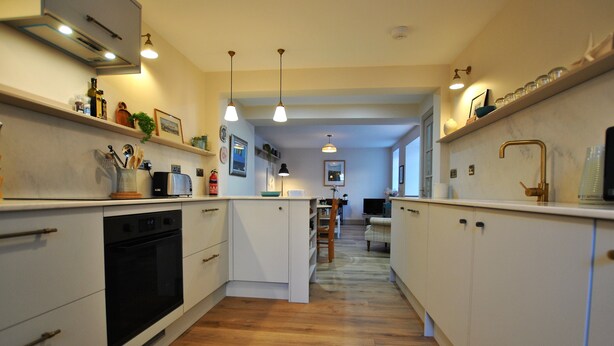 A narrow kitchen with white cabinets, a black oven, and wooden flooring. There are hanging pendant lights and a shelf with kitchen items. - Sea Holly Cottage- coastal retreat Pittenweem (Pittenweem)