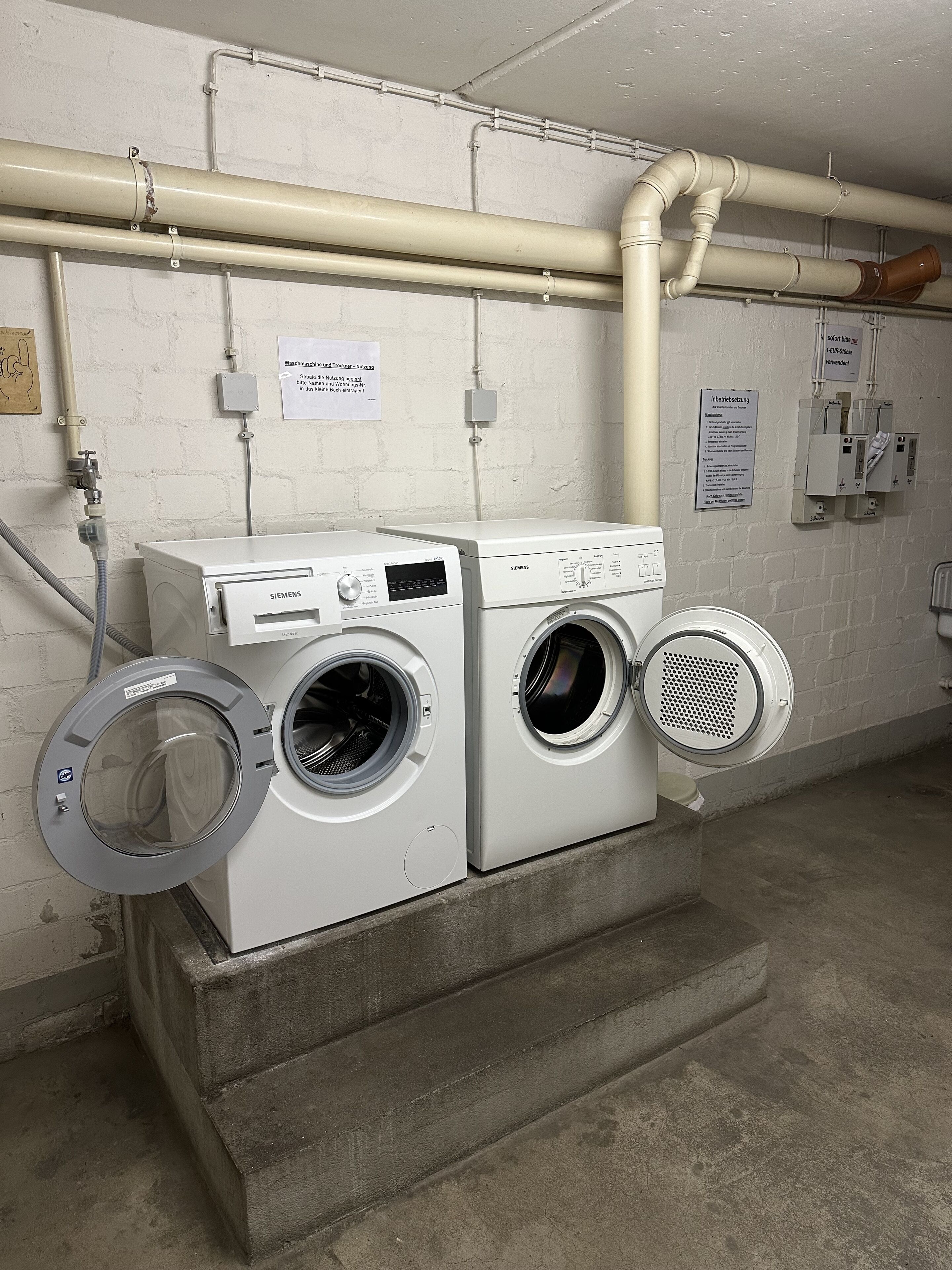 Eine Wäscherei mit zwei weißen Siemens Waschmaschinen, einer Wasserleitung und einem Bedienfeld an der Wand.