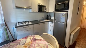 A compact kitchen with a gas stove, microwave, and refrigerator. There is a table with a patterned tablecloth and a chair.