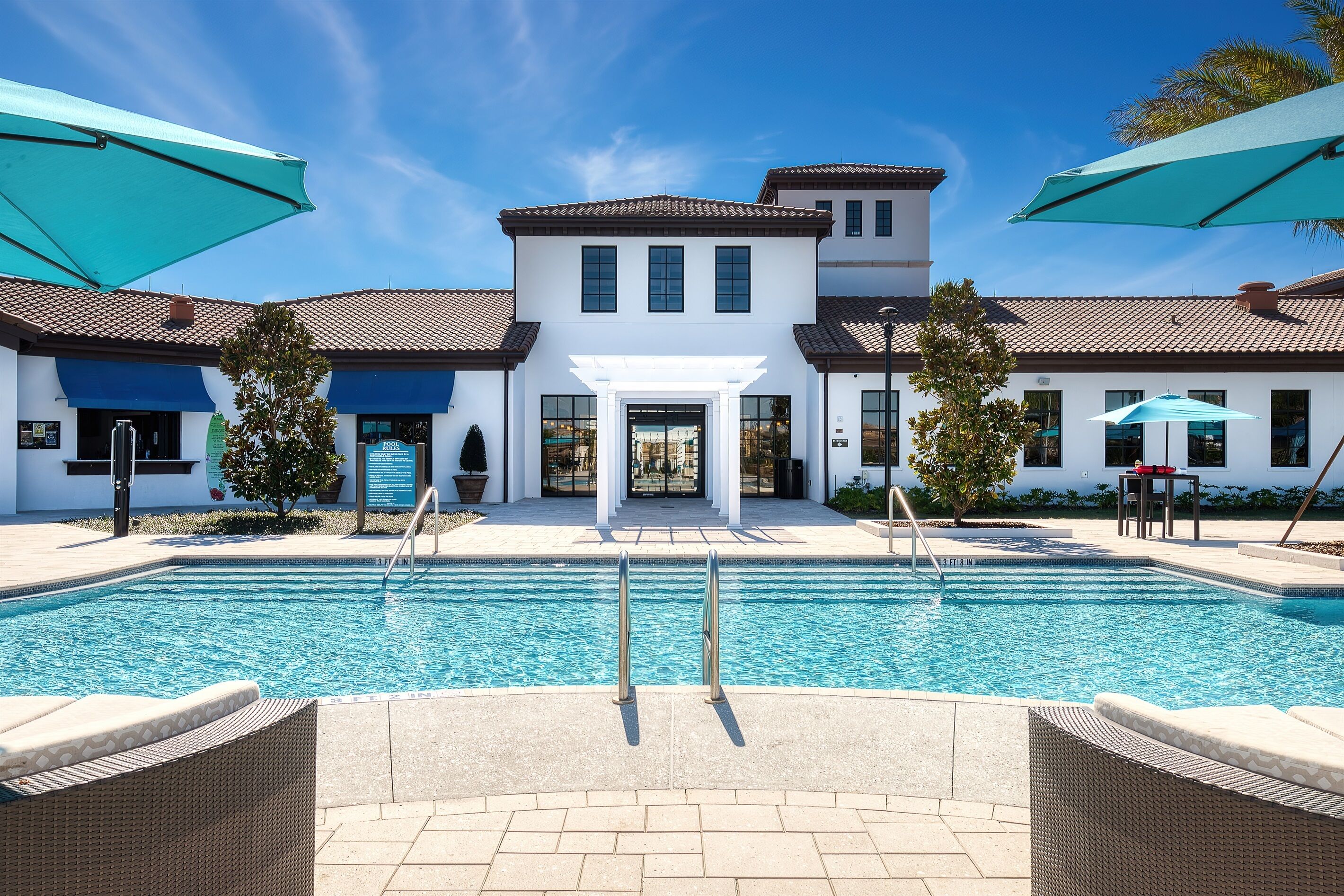 A swimming pool with a white building in the background, a covered patio area, and a clear blue sky.