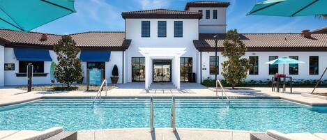 A swimming pool with a white building in the background, a covered patio area, and a clear blue sky.