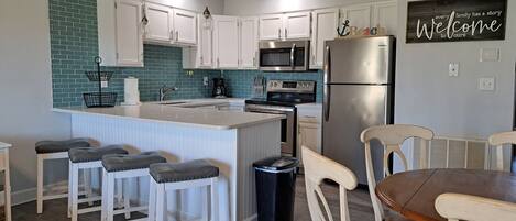 Una cocina moderna con armarios blancos, una isla central con taburetes altos y electrodomésticos de acero inoxidable, incluyendo un refrigerador y un microondas.