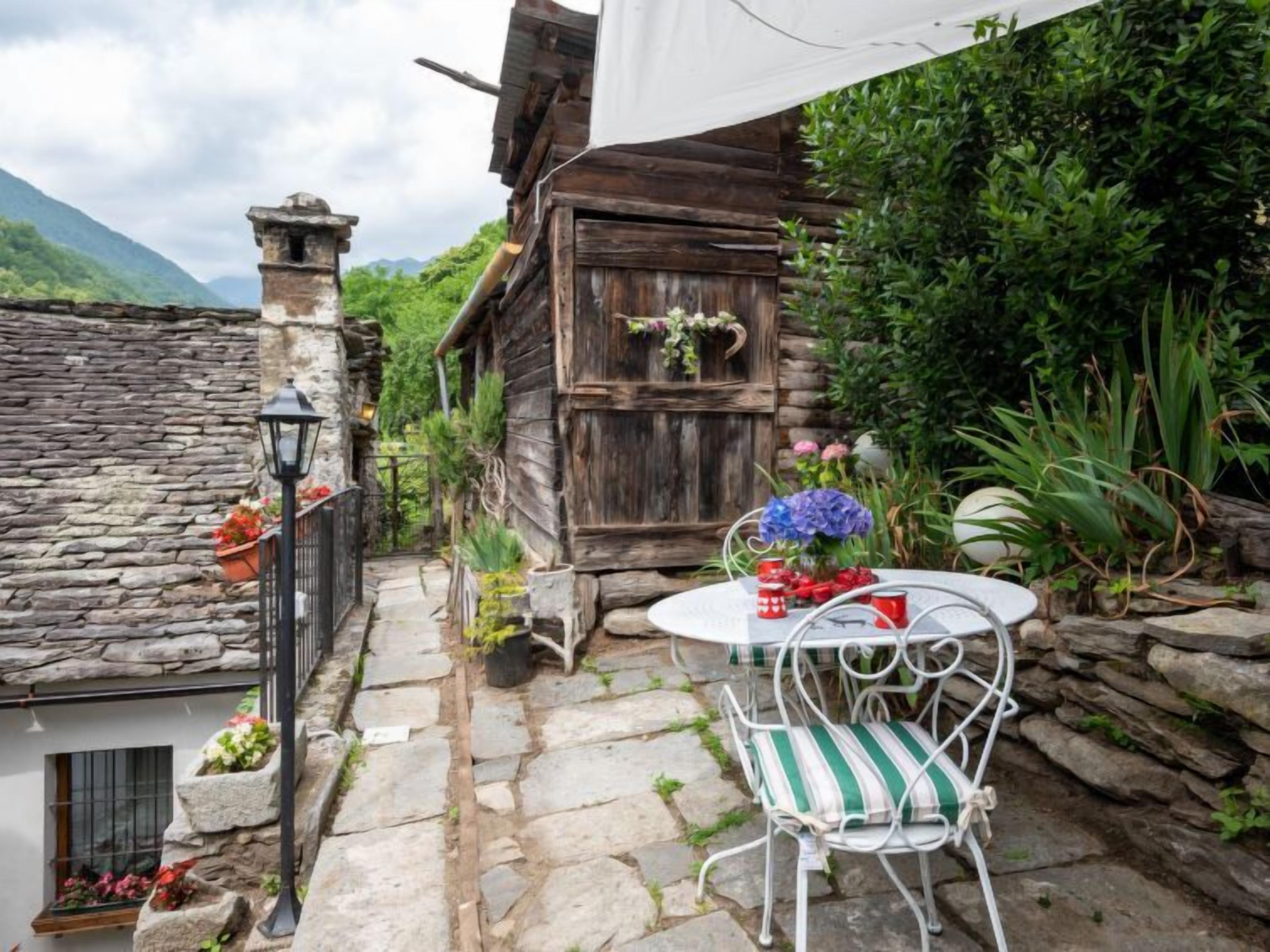 A rustic stone pathway leading to a wooden building with a chimney, a stone wall, and a small table with a chair.