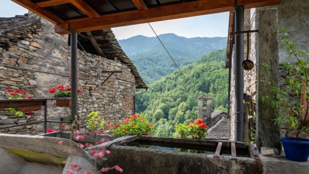 Un bâtiment en pierre surmonté d’un toit en bois, entouré d’une végétation luxuriante et avec des montagnes en arrière-plan.