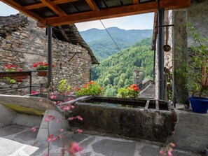Un bâtiment en pierre surmonté d’un toit en bois, entouré d’une végétation luxuriante et avec des montagnes en arrière-plan.