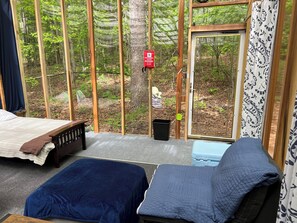 Ein Schlafzimmer mit einem Bett, zwei Kissen, einem kleinen Tisch und Blick auf einen Wald durch eine Glastür.