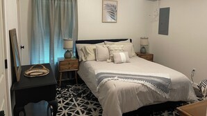 Une chambre à coucher comprenant un lit, deux tables de chevet, un ventilateur de plafond, une lampe fixée au mur, une œuvre d’art encadrée et une porte.