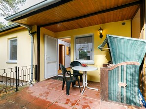 Ein überdachter Patio-Bereich mit Tisch und Stühlen, ein Korbsessel und eine Tür, die in einen anderen Raum führt.