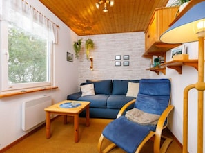 Una sala acogedora con un sofá azul, un sillón de madera mecedora, una mesita de madera y una ventana con cortinas.