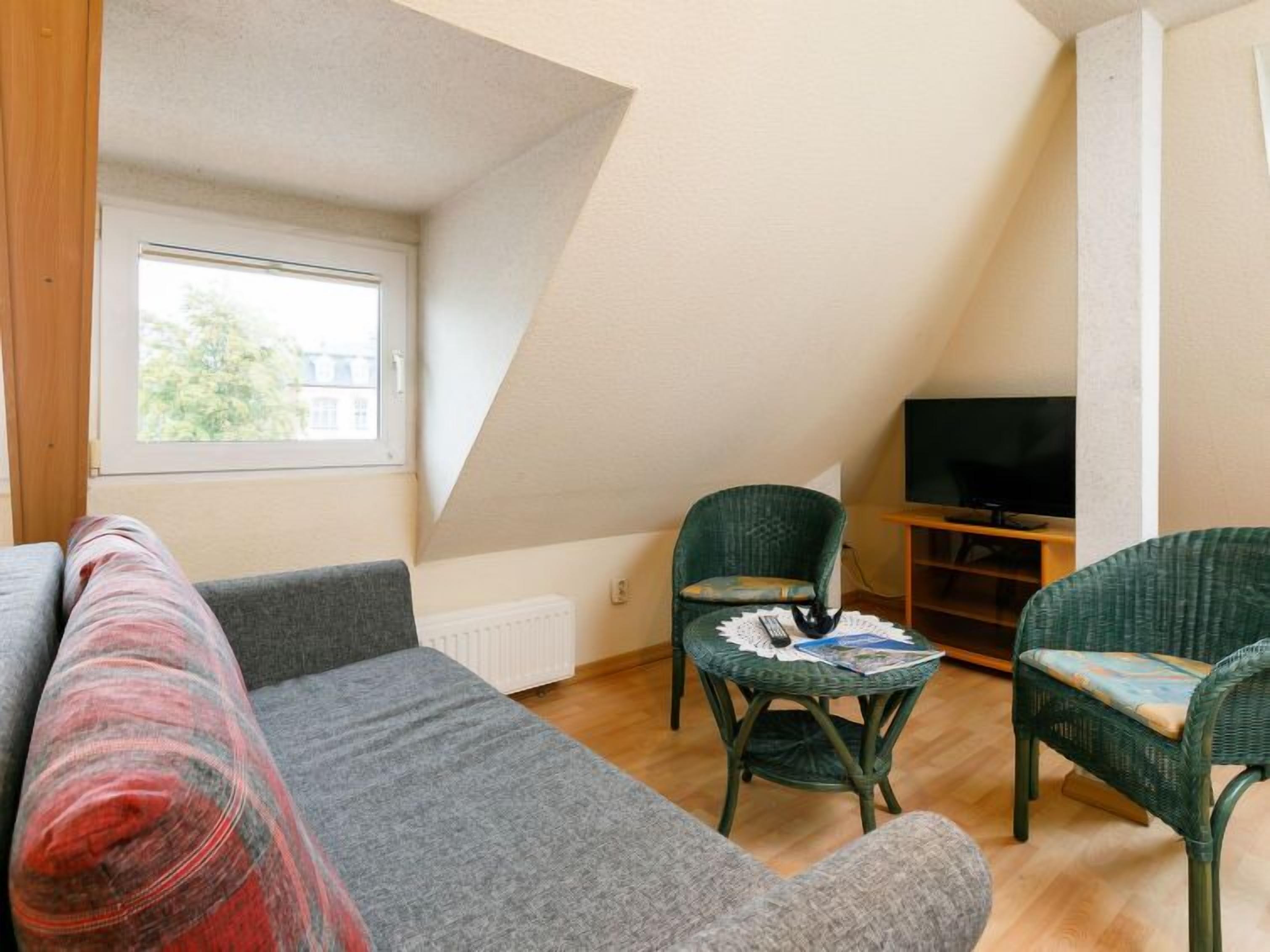 Un salon chaleureux avec un canapé, deux fauteuils en osier verts, une petite table avec une télécommande et un téléviseur posé sur un meuble en bois.