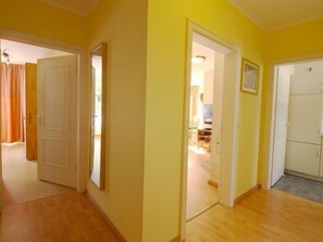 A hallway with wooden flooring and yellow walls, leading to a room with a television and a framed picture on the wall. - Luise von Preußen (App.101) by Interhome (Zinnowitz)