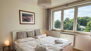 A neatly made bed with white linens and striped pillows, a bedside table with a lamp, a framed picture on the wall, and a window with a view of greenery. - Karsten by Interhome (Zinnowitz)