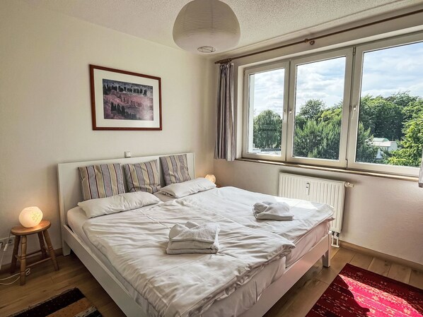 En pænt redt seng med hvide sengetøj og stribede puder, et natbord med en lampe, et indrammet billede på væggen og et vindue med udsigt over grøn vegetation.