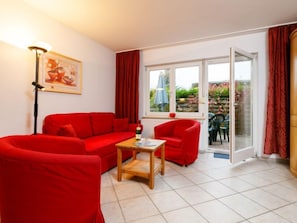 A living room with a red sofa, a wooden coffee table, and a red armchair. There is a painting on the wall and a window with a view of the garden. - Vacation home Susi by Interhome (Koserow)