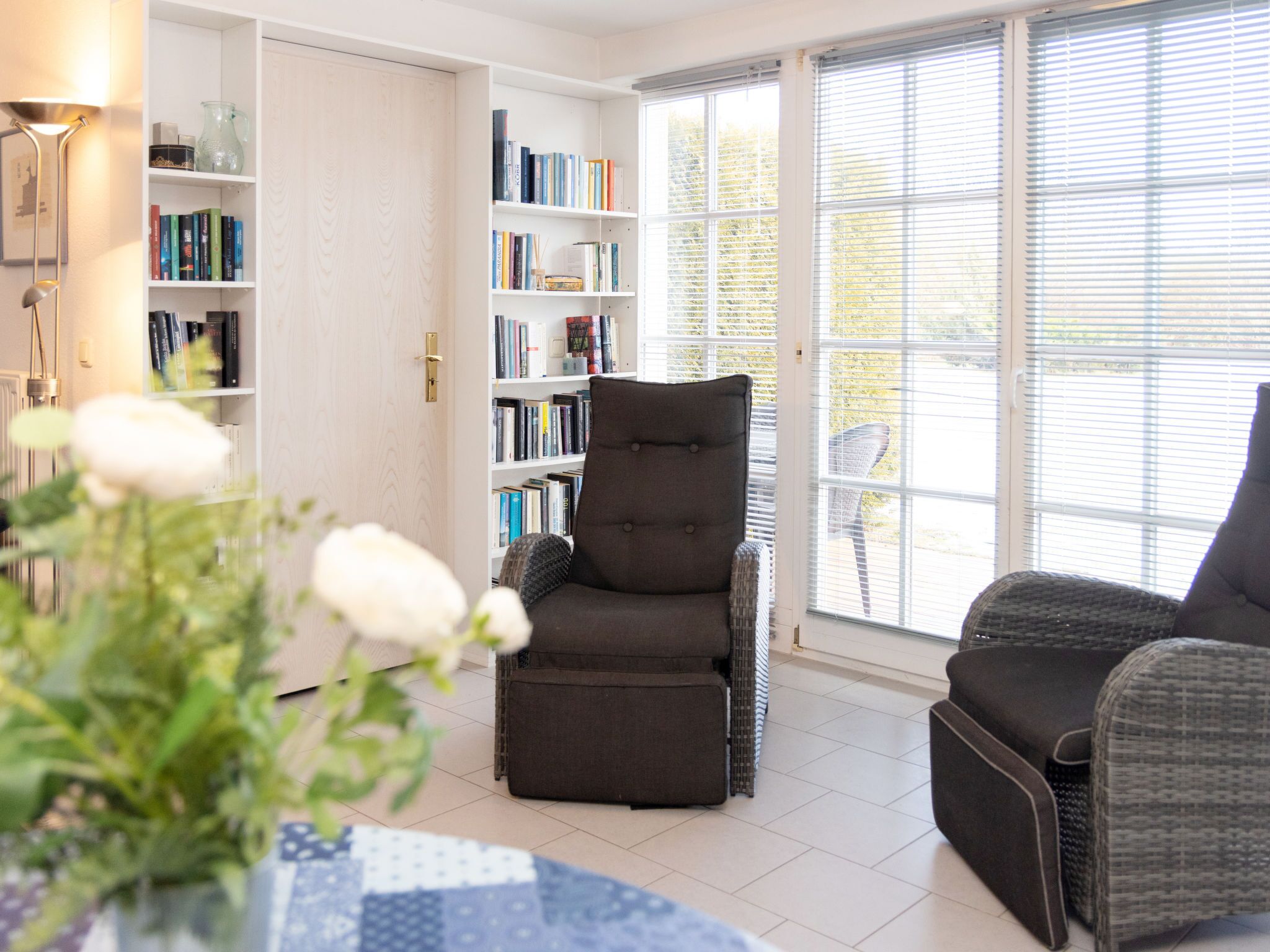 Ein Zimmer mit großem Fenster, einer Bücherwand, einem grauen Sessel und einem Tisch mit Blumen.