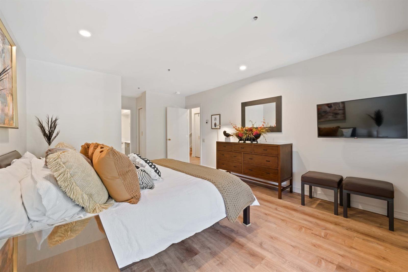 A spacious bedroom with a large bed, a wooden dresser, a bench, and a mirror on the wall.