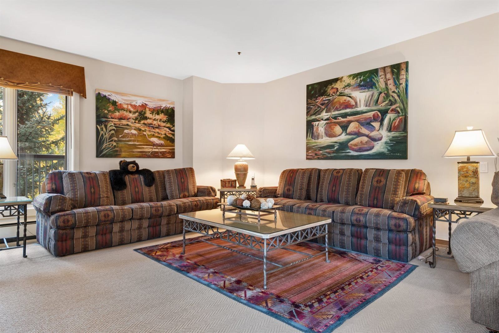 A living room with two sofas, a coffee table, and two lamps. There are two paintings on the walls and a window with a view of trees.