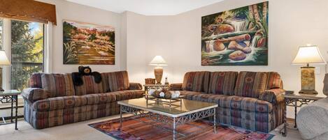 A living room with two sofas, a coffee table, and two lamps. There are two paintings on the walls and a window with a view of trees.