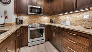 A modern kitchen with dark wooden cabinets, stainless steel appliances, a brick backsplash, and a granite countertop. - Beaver Creek Centennial 2 Bedroom #17 (Avon)