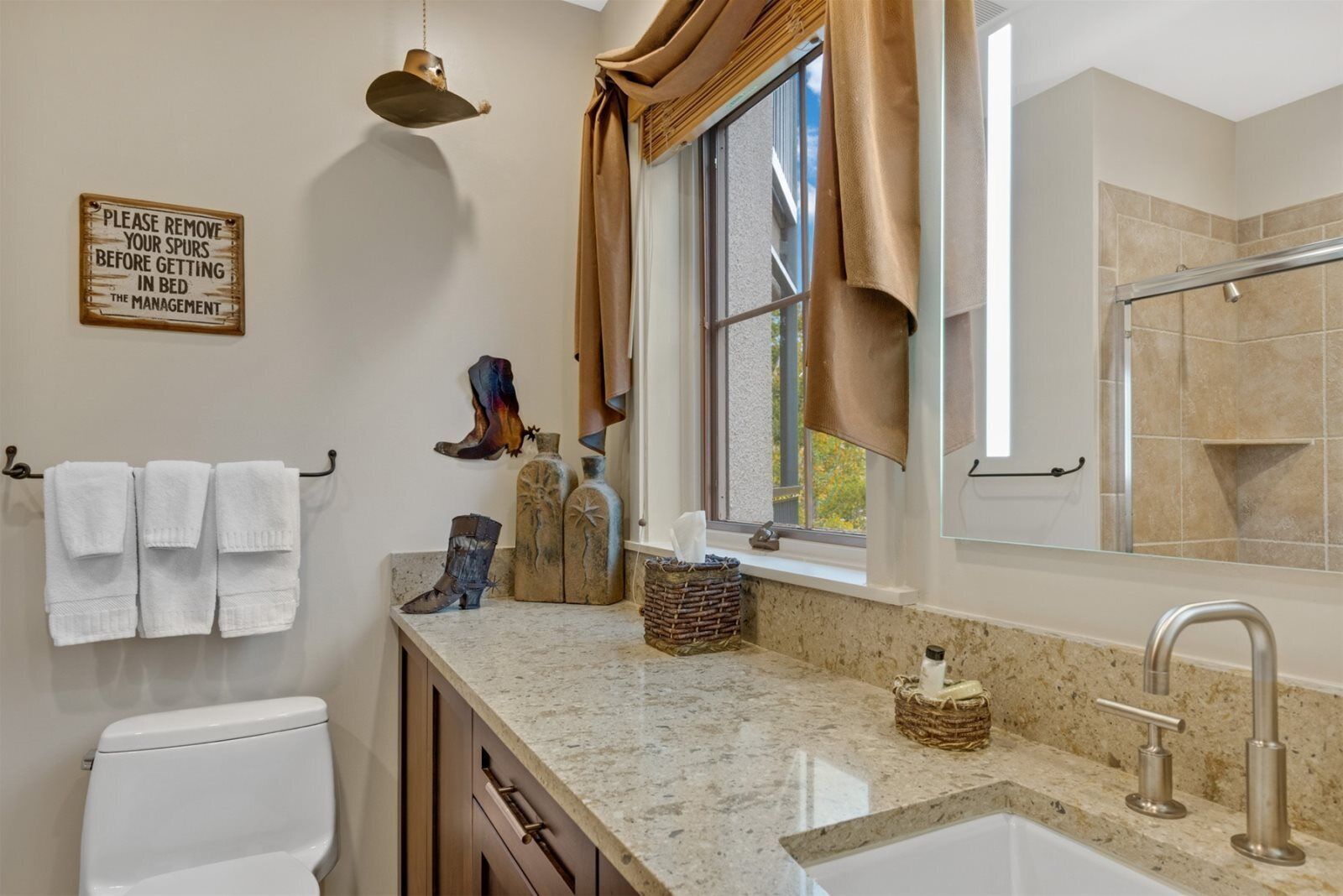 Baño con inodoro, lavabo y ventana con cortinas. Hay un letrero en la pared y un perchero con toallas.
