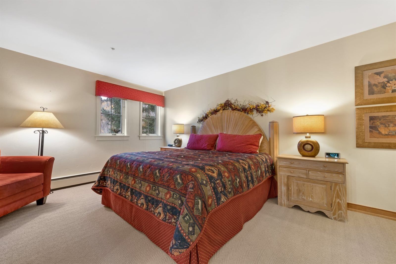 A bedroom with a large bed, two bedside lamps, a wooden nightstand, a red chair, and a window with a red valance.