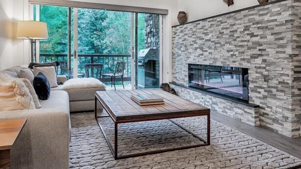 A modern living room with a stone fireplace, a sofa, a coffee table, and a view of the outdoors through large windows. - Beaver Creek Village Ski In, Ski Out 4 Bedroom (Avon)