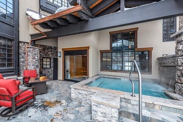 Un área de piscina moderna al aire libre con piso de piedra, un jacuzzi y asientos rojos.