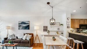 A modern living room with a sofa, coffee table, and dining area. There is a painting on the wall, a lamp, and a kitchen area with cabinets and a countertop. - Lakeside Village 1 Bedroom Condo in Lakeshore Complex (Keystone)