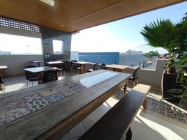 Uma área de refeições ao ar livre com uma mesa de madeira comprida e várias cadeiras, rodeada por plantas em vasos e vista para a cidade.
