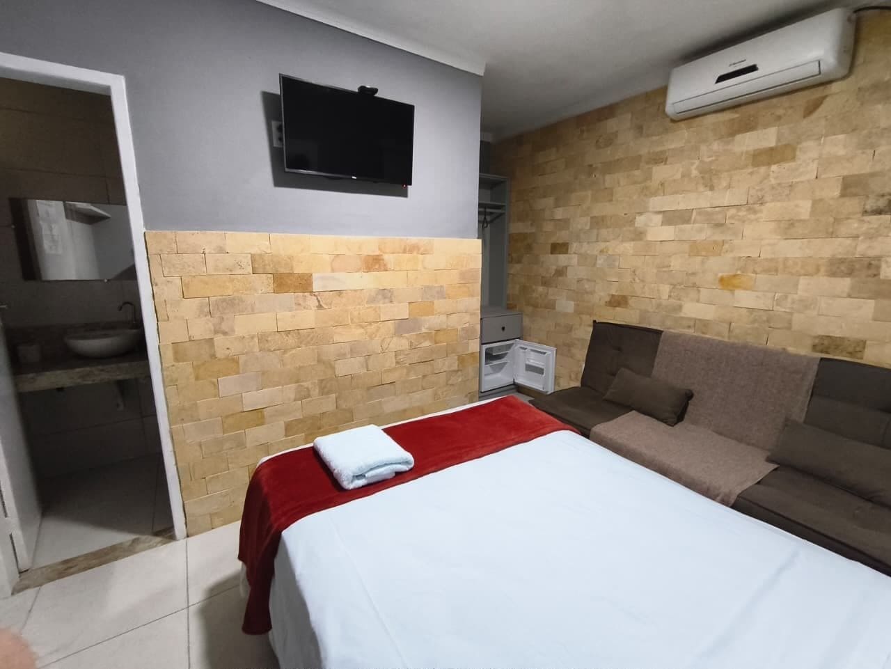 Quarto de hotel com uma cama, uma manta vermelha, um sofá marrom, uma televisão e um aparelho de ar condicionado.