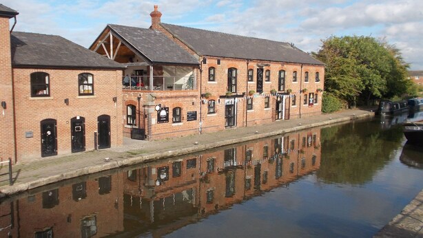 A brick building with a balcony, a canal, and a boat docked on the right. - Idyllic Bungalow, Ideal Location in West Lancashire (Burscough)